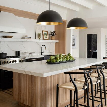 A wide view into the kitchen across a large island countertop to the stove and sink on the far wall. Two large pendant lamps hang painted with black exterior and reflective gold interior above the white marble island.